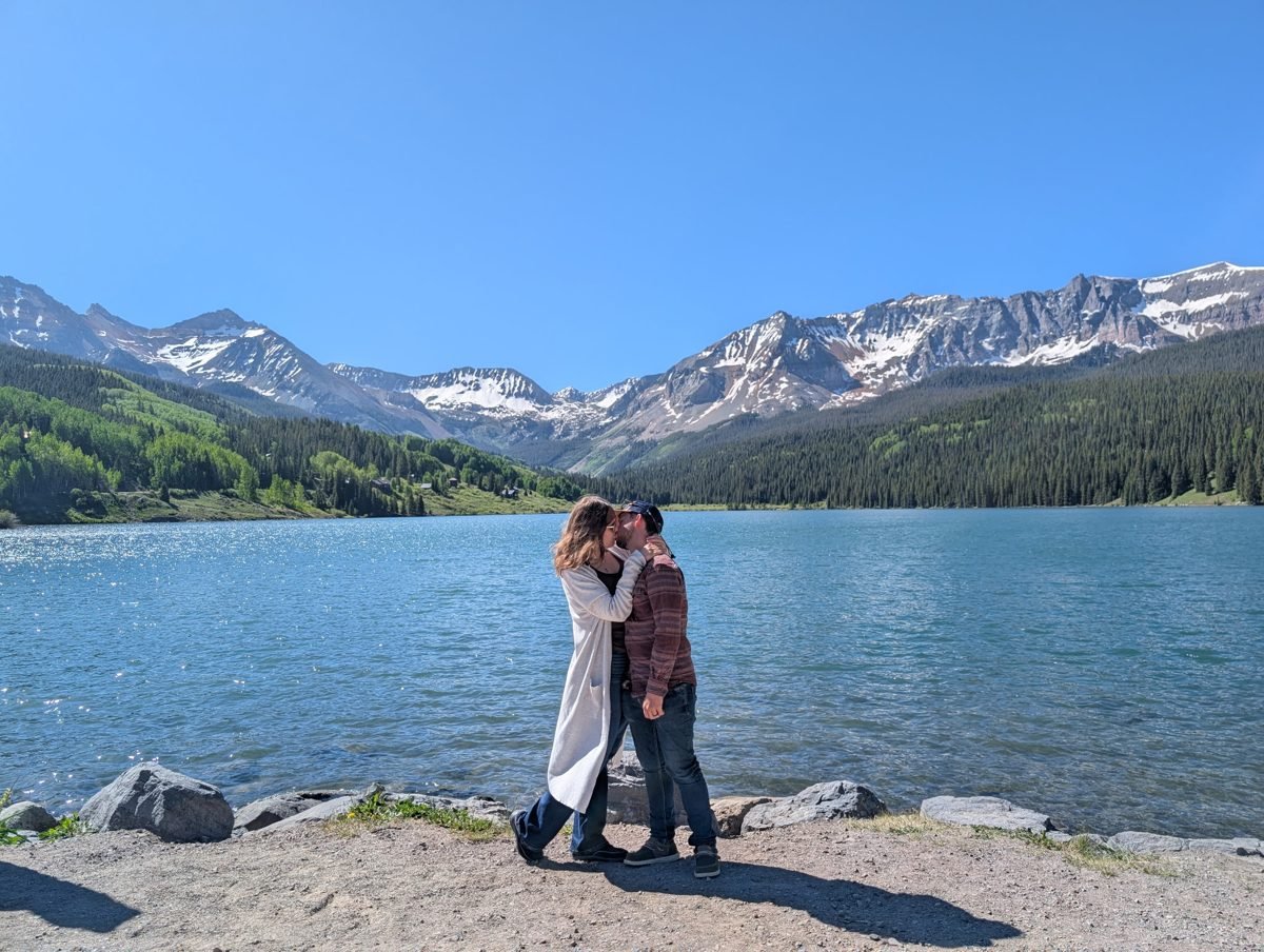 Margaret and her fiancé kissing on the shore of a mountain lake, snow-capped peaks behind them.